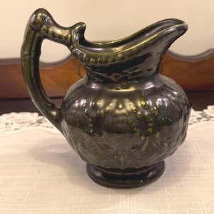 May include: A dark green ceramic pitcher with a bulbous body and a curved handle. The pitcher has a spout for pouring and sits on a small, round base. The surface has a textured, mottled appearance.