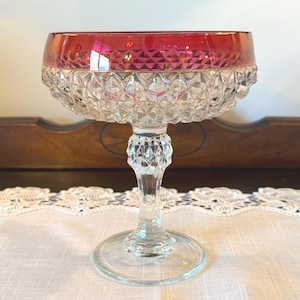 May include: A vintage champagne coupe glass with a ruby red rim and a clear, diamond-cut bowl. The glass sits on a clear stem and base, showcasing a classic design. The glass is likely made of crystal.