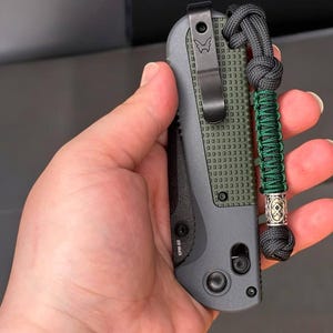 Puede incluir: Un cuchillo de bolsillo negro y verde con un mango texturizado y un clip. El cuchillo tiene un cordón con un cordón trenzado verde y negro y una cuenta de plata.