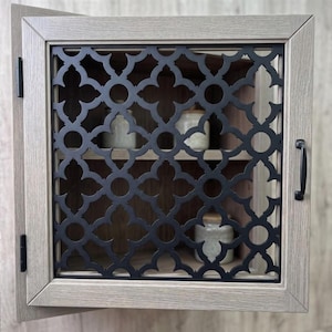 Puede incluir: Un armario de pared cuadrado con acabado de veta de madera clara y una puerta de metal negro con un patrón geométrico repetitivo de cuatrifolio. El armario tiene dos bisagras negras en el lado izquierdo y un tirador negro a la derecha.