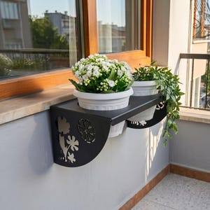 May include: A black metal wall-mounted planter shelf featuring decorative cut-out flower patterns on the side panels. The shelf holds two white plastic pots, one containing a white flowering plant and the other a trailing green leafy plant.