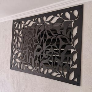 Può includere: Una griglia di ventilazione rettangolare in metallo nero con un motivo tagliato al laser di forme di foglie organiche. Il pannello decorativo è montato sopra una bocchetta d'aria a lamelle orizzontali e fissato con viti.