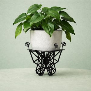 May include: A potted plant with large green leaves sits in a speckled white pot. The pot rests on a black metal plant stand with a geometric design and flower-shaped accents. The stand is on a light green background.