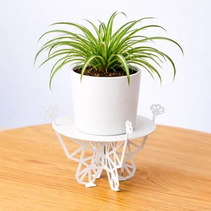 Op de afbeelding: Een witte keramische pot met een groene plant staat op een witte metalen plantenstandaard. De plantenstandaard heeft een geometrisch ontwerp en een ronde bovenkant. De plant heeft lange, groen-wit gestreepte bladeren. De standaard staat op een houten oppervlak.