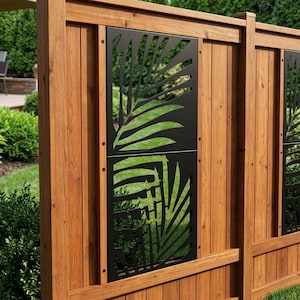 Op de afbeelding: Een houten schuttingpaneel met een verticaal rechthoekig zwart metalen inzetstuk met een lasergesneden palmbladpatroon. De schutting is opgebouwd uit verticale bruine houten planken die worden omlijst door horizontale rails met zwarte schroeven.