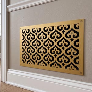 May include: A rectangular gold-colored metal vent cover featuring a repeating intricate cutout pattern of stylized fleur-de-lis shapes. The cover is mounted flush against a wall with four visible screws, one in each corner of the frame.