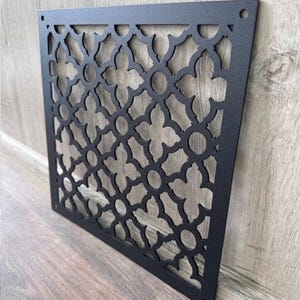 May include: A square, matte black metal vent cover featuring a repeating quatrefoil and circular cutout pattern. The flat, rigid panel includes small circular mounting holes in the top corners, designed for installation over an air duct opening.