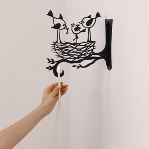 May include: Black metal wall art featuring a bird family in a nest on a tree branch. The design includes three birds, a nest, and leaves. A white hook is attached to the branch, suitable for hanging items. The art is mounted on a white wall.