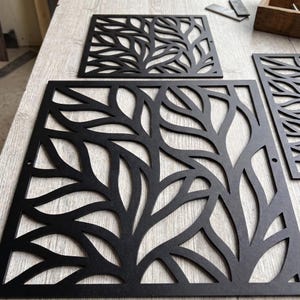 May include: A set of square black metal panels featuring a laser-cut leaf pattern. Each panel has a frame and a repeating organic foliage design with cutouts. The panels are flat and have small circular holes on the sides for mounting purposes.