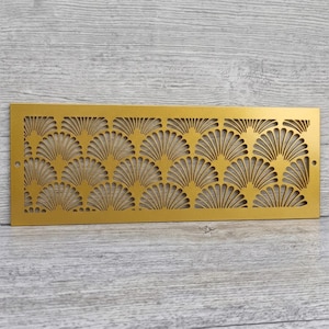 May include: A rectangular gold-colored metal vent cover featuring a repeating fan-shaped cutout pattern. The flat panel has a smooth finish and includes a small circular screw hole on both the left and right sides for mounting purposes.