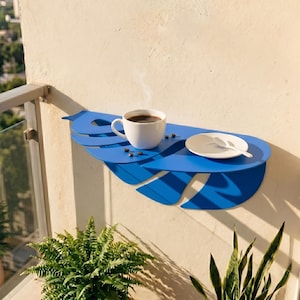 Puede incluir: Un estante de pared azul brillante con forma de hoja tropical grande. El estante sostiene una taza de café de cerámica blanca con líquido oscuro humeante, un platillo blanco, una cuchara blanca y varios granos de café tostados dispersos.