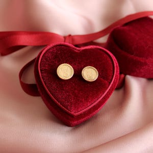 May include: A pair of gold-colored stud earrings with a portrait design, displayed in a heart-shaped, red velvet jewelry box. The box is open, revealing the earrings, and sits on a pink satin surface. A red ribbon is tied around the box.