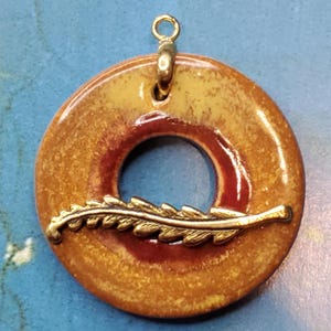 May include: A round ceramic pendant with a gold leaf design. The pendant is a light brown color with a red ring around the center hole. The pendant has a gold bail for hanging.