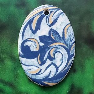 May include: Oval-shaped ceramic pendant with a floral design. The pendant features a white base with blue and gold-colored floral patterns. A small hole is at the top for hanging. The edges are white, and the background is green.