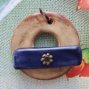 May include: A ceramic pendant with a beige and brown ring shape with a blue rectangular piece in the center. The blue piece has a decorative metal flower with a brown center.
