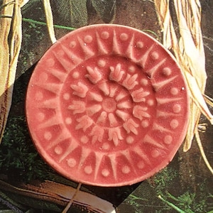 May include: A round, pink ceramic coaster with a raised, intricate design. The design features a circular pattern of repeating floral shapes.