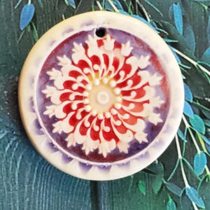 May include: A round, ceramic pendant with a floral design. The pendant features a central cream-colored circle with a red and white floral pattern. The outer edge is cream-colored with a purple border. A small hole is at the top for hanging.