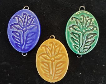 Op de afbeelding: Drie ovale hangers van keramiek met een bloemenmotief. De hangers zijn blauw, groen en geel.
