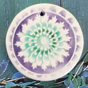 May include: A round ceramic ornament with a white, green, and purple mandala design. The ornament has a hole at the top for hanging.