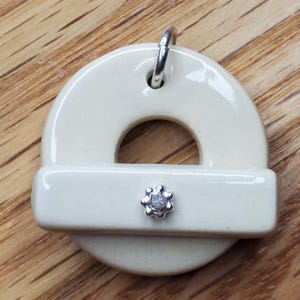 May include: A white ceramic pendant with a silver ring at the top and a silver star-shaped charm in the center. The pendant is shaped like a circle with a hole in the middle.