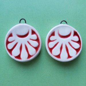 May include: Two white ceramic pendants with a red floral design. Each pendant has a small, dark metal loop at the top for hanging. The pendants are round and have a raised, textured pattern.