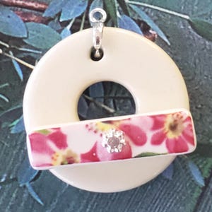 May include: Cream-colored pendant with a circular design and a rectangular floral accent. The accent features pink and red flowers with a silver-colored gem in the center. A silver-colored bail is attached at the top.