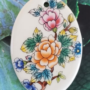 May include: Oval ceramic pendant with a floral design. The pendant features a white background with a branch of flowers in shades of pink, orange, yellow, blue, and green.