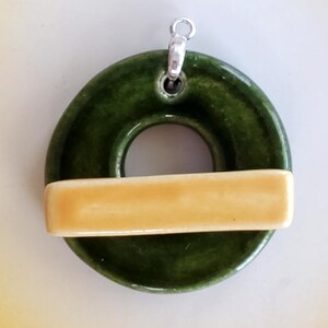 May include: A green ceramic pendant with a yellow band across the center. The pendant has a hole in the center and a silver loop at the top for hanging.