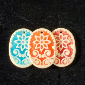 May include: Three ceramic ornaments in the shape of an oval with a white floral design. The ornaments are painted in shades of blue, orange, and red.
