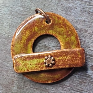 May include: A brown and green ceramic pendant with a ring design and a smaller bar attached. The bar has a decorative metal piece in the center.