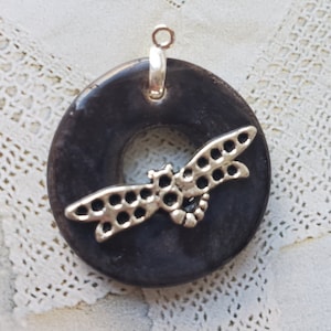 May include: A black ceramic pendant with a silver dragonfly design. The pendant has a hole in the center and a silver loop for hanging.