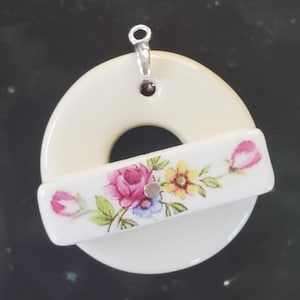 May include: A white ceramic pendant with a floral design. The pendant is shaped like a ring with a floral design on a rectangular piece that is attached to the ring.