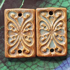 May include: Two rectangular ceramic tiles with a brown glaze and a light blue glaze. The tiles have a decorative design of a stylized flower with a hole in the center.
