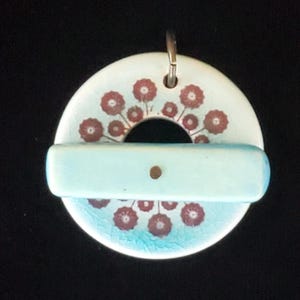 May include: A ceramic pendant with a circular top and a rectangular bar. The top is white with a floral pattern in red and a blue cracked glaze. The bar is light blue with a small hole. A silver-colored bail is attached.