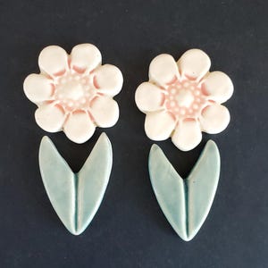 May include: Two ceramic flower and leaf set. The flowers are white with pink centers, and the leaves are a light blue-green color. The set is arranged on a dark background. The flower petals are rounded.