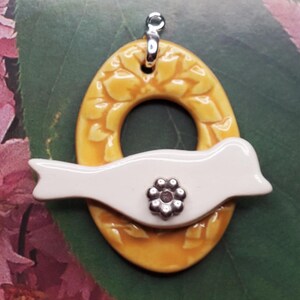 May include: A ceramic pendant featuring a white bird with a silver flower detail, perched on a yellow oval with a floral design.