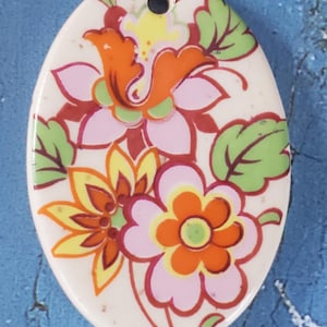 May include: Oval ceramic pendant with a floral design. The pendant features pink, orange, yellow, and green flowers on a white background.