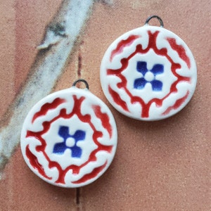 May include: Two white ceramic round pendants with red and blue floral designs. The pendants have a hole at the top for hanging.
