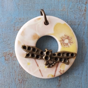 May include: A white ceramic pendant with a floral design and a bronze dragonfly charm. The pendant is shaped like a donut with a hole in the center.