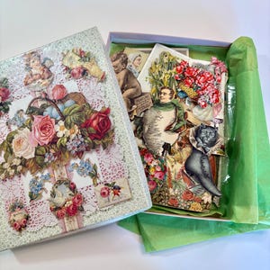May include: A rectangular box with a lid featuring a collage of vintage floral illustrations, roses, and cherubs. Inside, the box contains a collection of assorted paper ephemera, including Victorian-style cutouts of flowers, a cat, and figures, lined with bright green tissue paper.