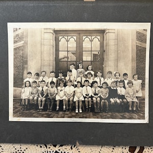 May include: A vintage black and white photograph of a large group of children posed in rows in front of a brick building with double doors. The children are dressed in formal attire, including collared shirts, ties, and dresses, arranged in three tiers.