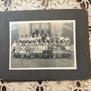 May include: A black and white vintage photograph mounted on a dark rectangular board, depicting a large group of children posed in rows in front of a brick building with double doors. The children are dressed in formal attire, including collared shirts.