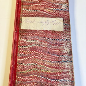 May include: A rectangular antique book with a red spine and a marbled cover featuring a repeating pattern of red, yellow, blue, and white swirls. A white paper label is affixed to the center with the handwritten text May 12 - July 9th 1880.