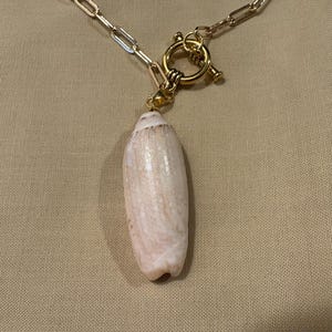 May include: A gold-toned chain necklace with a toggle clasp and a pendant made from a light-colored seashell. The seashell pendant is elongated and has a natural, textured surface.