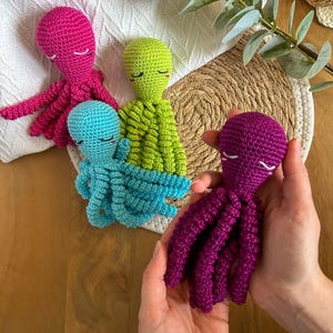 Peut inclure: Plusieurs jouets pieuvres crochetés à la main, de couleurs rose vif, vert citron et turquoise. Une personne tient une pieuvre violet foncé. Chaque pieuvre a les yeux fermés et huit tentacules. Les jouets sont sur une surface en bois.