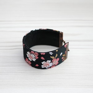 Puede incluir: Un brazalete de tela negro con un patrón floral en rosa y blanco. El brazalete tiene un cierre de cobre.