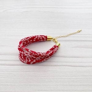 Puede incluir: Pulsera de tela roja con un patrón floral blanco y un cierre dorado.