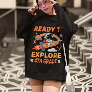 May include: Black sweatshirt with a colorful graphic of an astronaut on a rocket-pencil, a rainbow, and the words "READY TO EXPLORE 4TH GRADE". The design includes planets, stars, and clouds.
