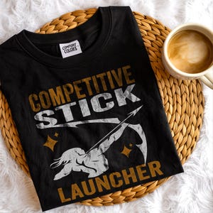 May include: Black t-shirt with the text "COMPETITIVE STICK LAUNCHER" in white and gold lettering. The shirt features a graphic of a person with a bow and arrow. The shirt is on a woven mat with a cup of coffee.