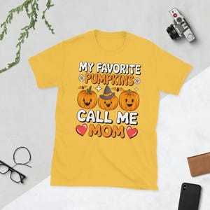 May include: A mustard yellow short-sleeve t-shirt featuring a graphic print of three orange pumpkins with faces. The center pumpkin wears a witch hat. The text reads MY FAVORITE PUMPKINS CALL ME MOM, accented with small flowers, bats, and red hearts.